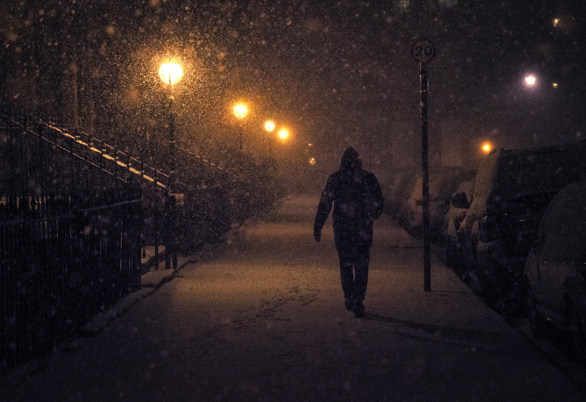 Person geht bei Schneefall am Abend durch dunkle von Laternen beleuchtete Straße