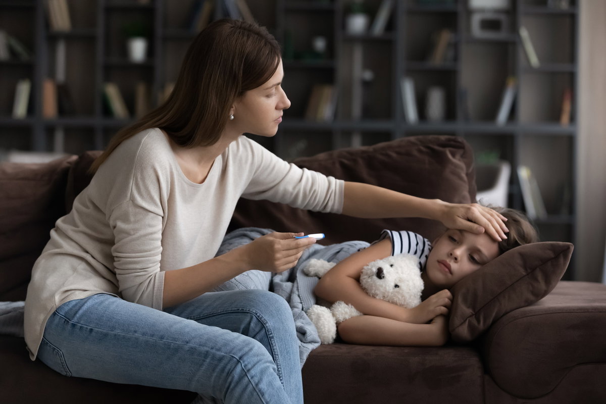 Kind liegt krank auf dem Sofa, während die Mama Fieber misst