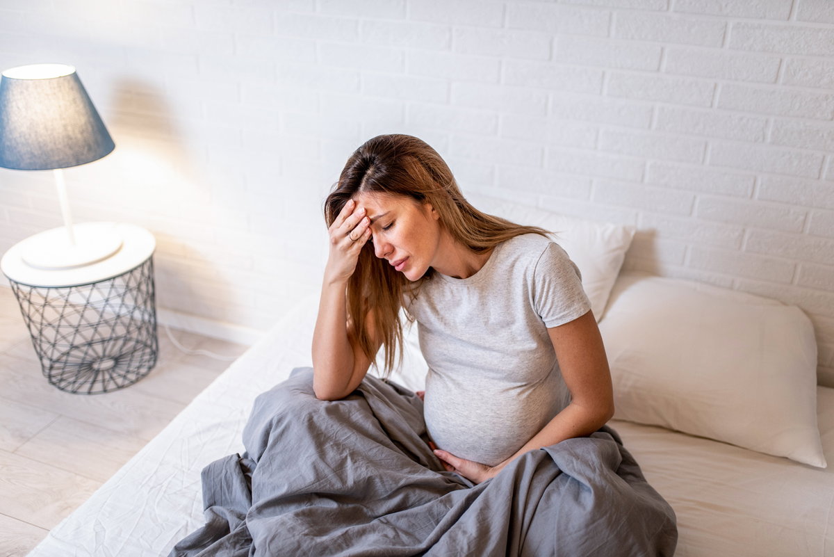 Schwangere sitzt im Bett und hält sich Kopf und Bauch