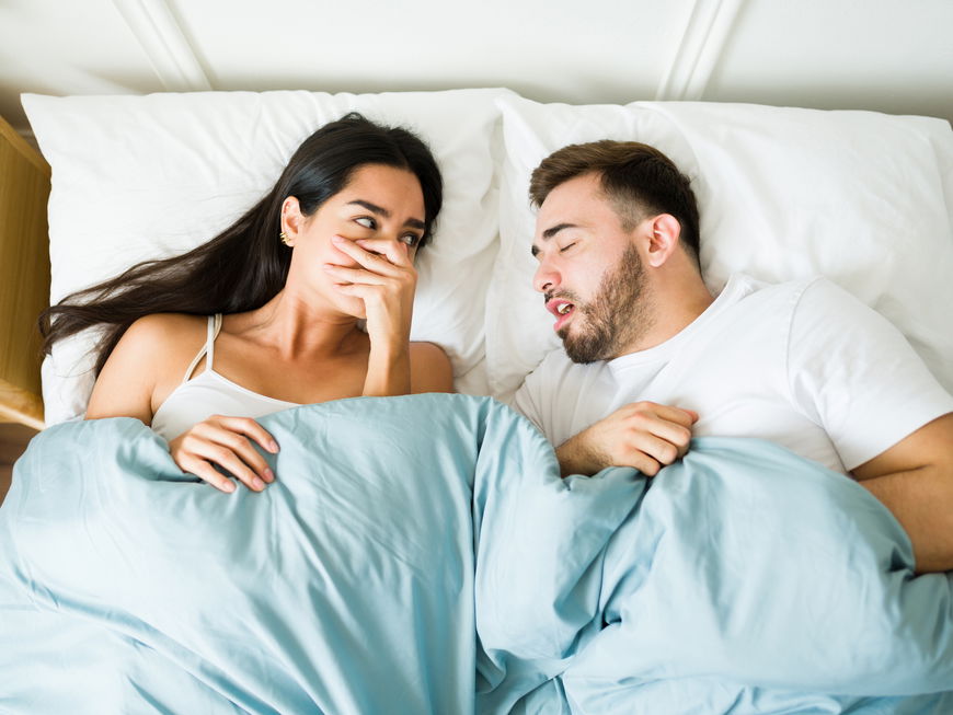 Frau liegt neben ihrem Freund im Bett und hält sich die Hand vor den Mund