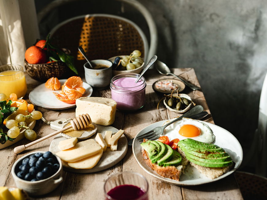 Bunt gedeckter Frühstückstisch mit Käse, Obst, Joghurt und Avocado-Brot mit Spiegelei
