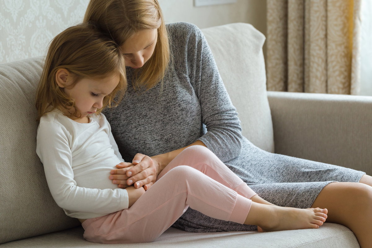 Bauchschmerzen bei Kindern: Mädchen sitzt mit Bauchschmerzen auf der Couch und wird von der Mutter getröstet.