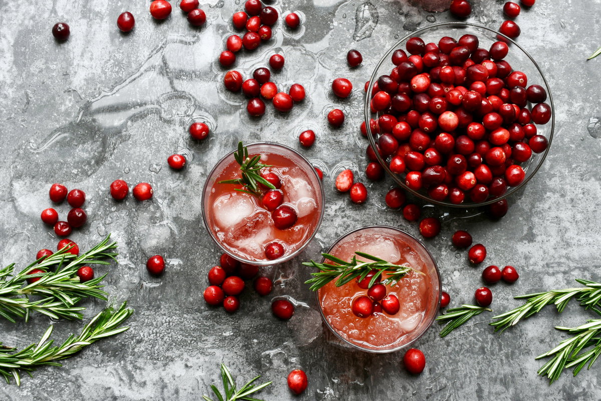 Draufsicht auf eine Schale mit frischen Cranberrys und zwei Gläsern Cranberrysaft mit Eiswürfeln und Rosmarin