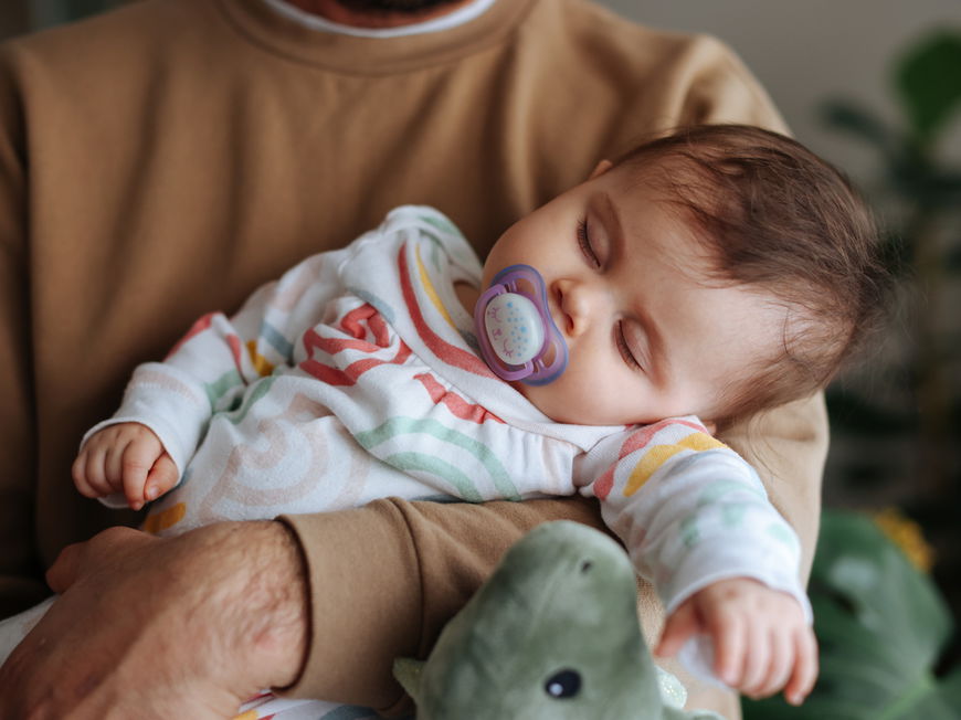 Ein Baby schläft mit einem Schnuller auf dem Arm des Vaters.
