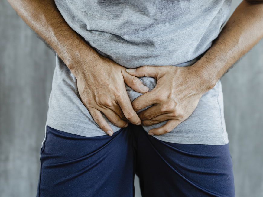 Nahaufnahme eines Mannes in kurzer blauer Hose mit grauem Shirt, der sich vor Schmerzen in den Schritt greift