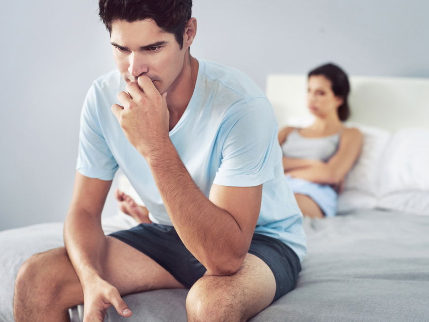 Mann sitzt auf dem Bett und ist verzweifelt mit Frau im Hintergrund