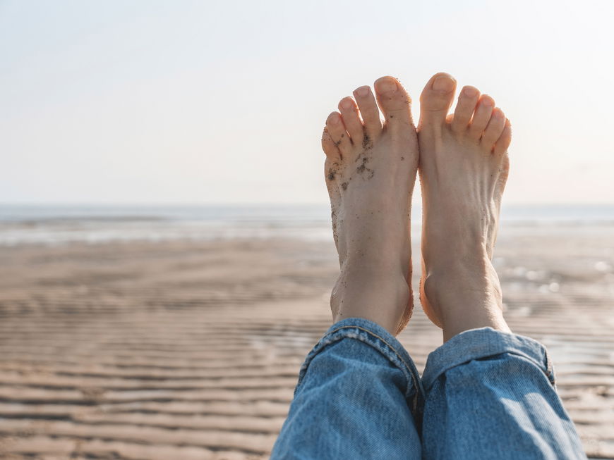 Eine Frau hält ihre nackten Füße hoch. Sie liegt am Strand. Ihre Fußnägel sind naturbelassen.