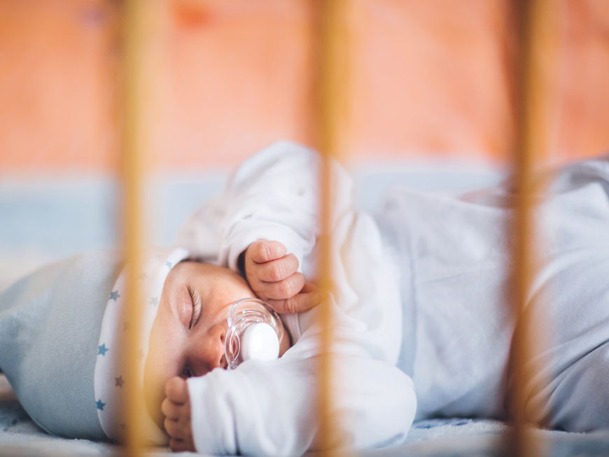 Baby schläft mit Schnuller und Mützchen im Gitterbett